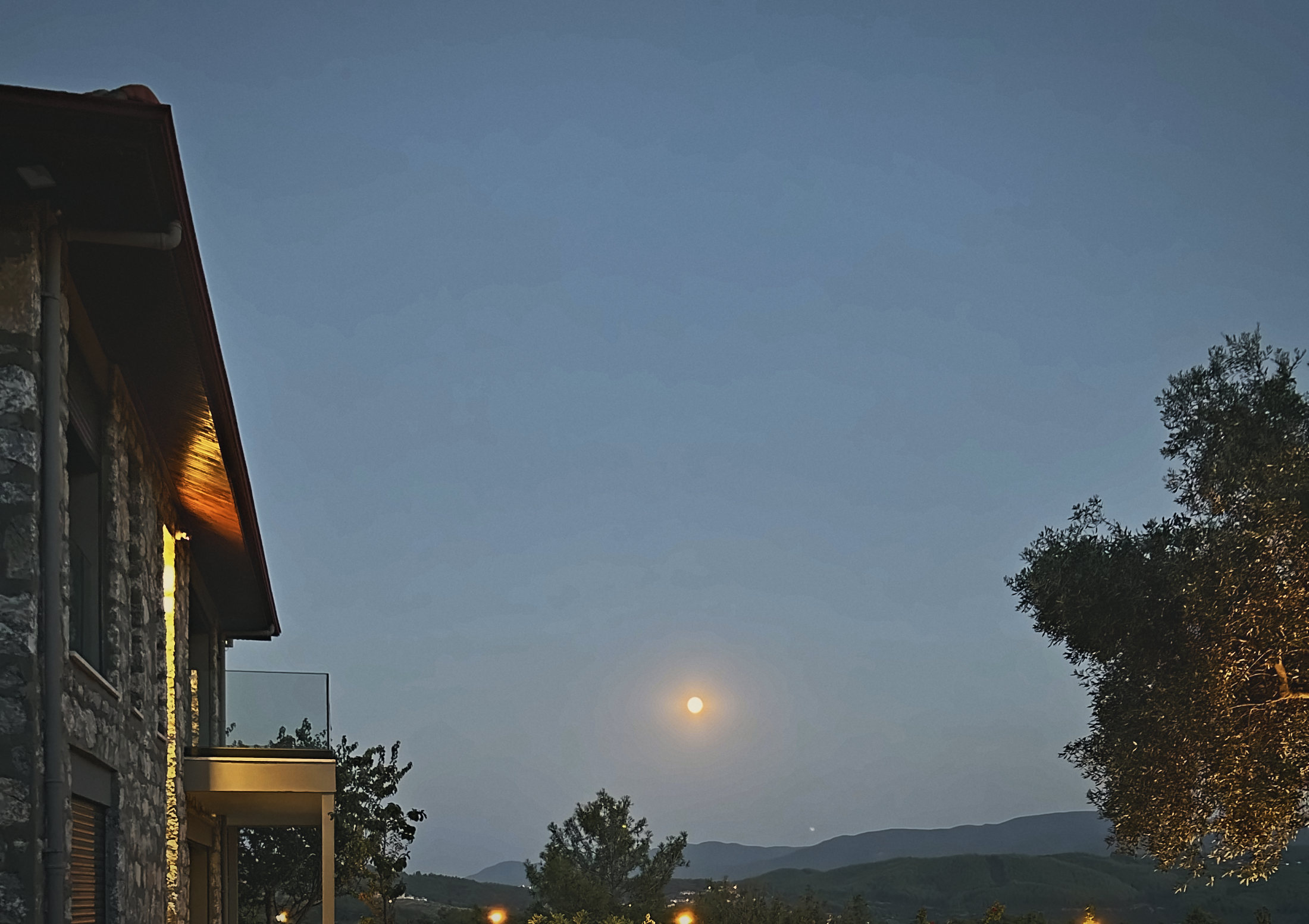 A full moon rising over the valley beside the lit stone house